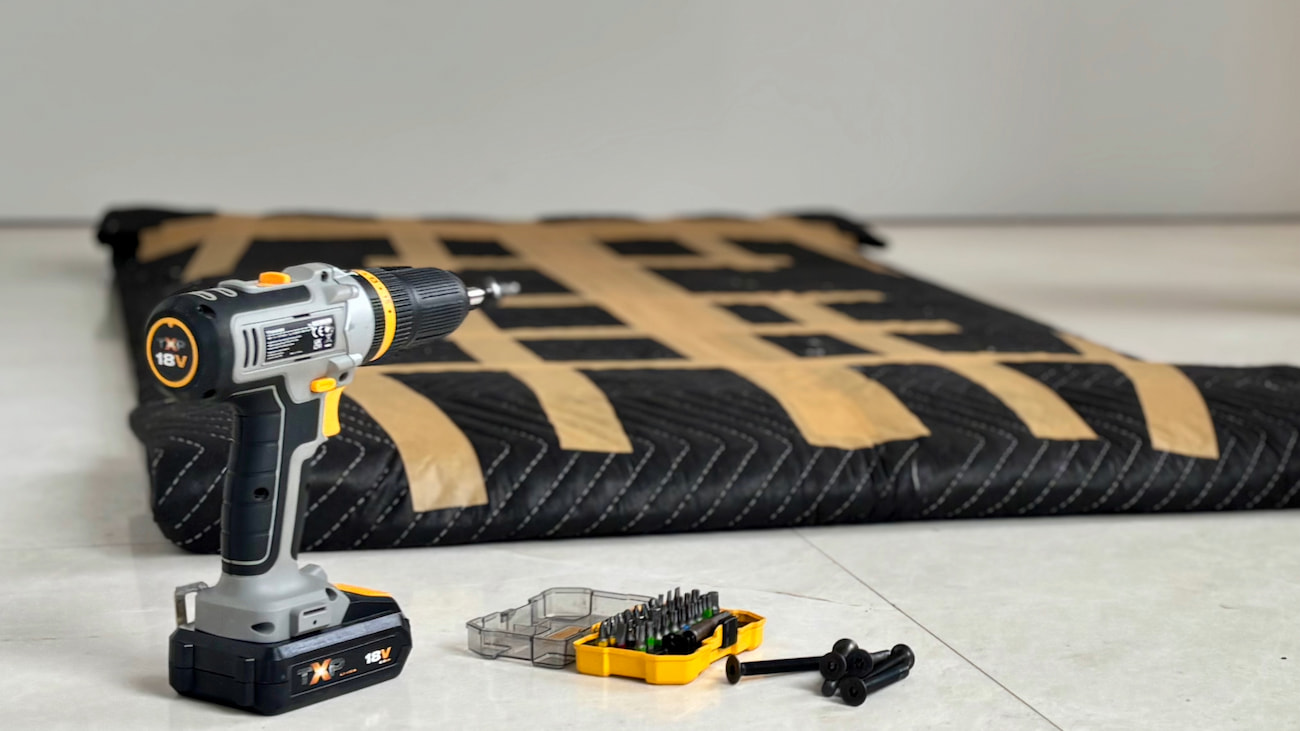 A wooden table wrapped securely in padded moving blankets next to a cordless power drill and bits for furniture disassembly.